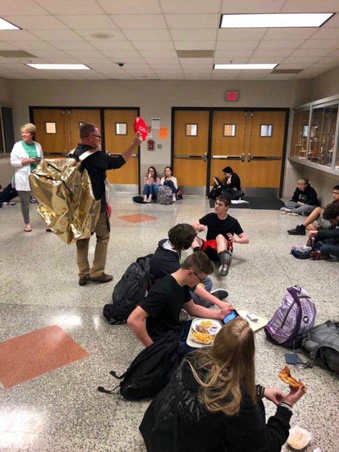 Mr. Jason Ruyle wears an oversized gold backpack while passing out water bottles, frisbees and t-shirts to students.